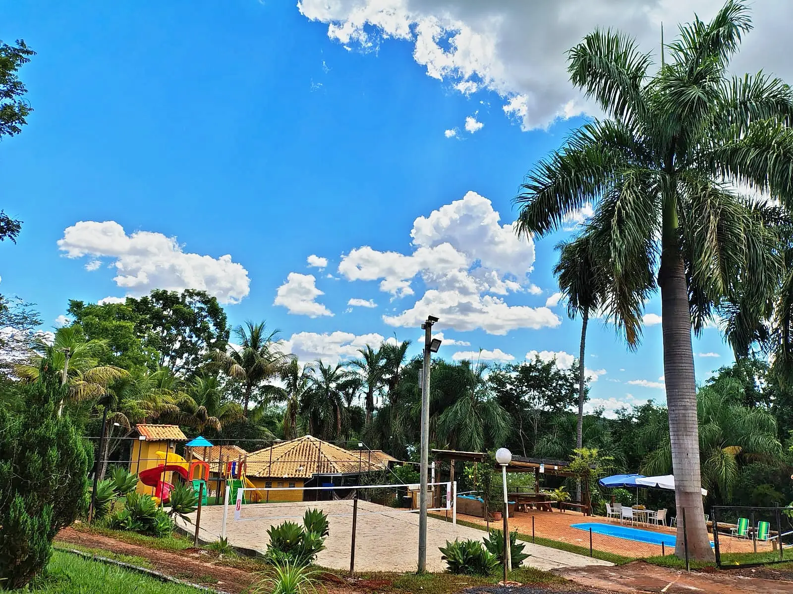 Vista da área de lazer do Rancho SPPA em dia de sol: quadra de areia, piscina, playground, quiosque e coqueiros